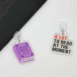 May include: Two clear acrylic charms with silver-colored hardware. One charm is a purple book with the text "I Read A Lot But I'm So Productive It's An Art". The other charm says "A LOT TO READ AT THE MOMENT" in red and black.
