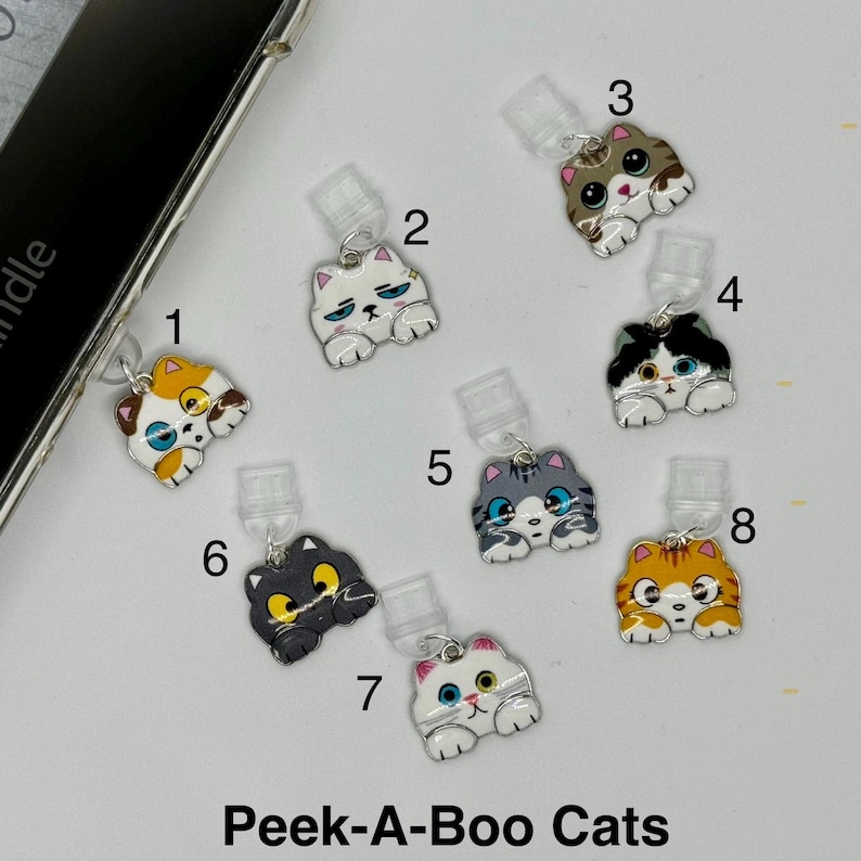May include: A collection of eight cat-shaped phone charms with various color schemes, including orange, gray, black, and white. Each charm has a silver-colored attachment. The text "Peek-A-Boo Cats" is displayed at the bottom.