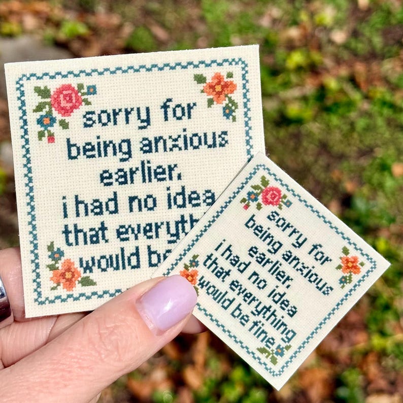 May include: Two square cross-stitch pieces on off-white fabric with dark blue text reading, "sorry for being anxious earlier. i had no idea that everything would be fine." The borders feature a dark blue grid pattern with pink and orange flowers.