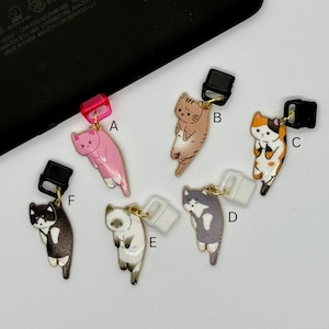 May include: A collection of six cat-shaped charms in various colors, including pink, brown, calico, gray, white, and black. Each charm has a gold-colored loop and a black or white connector. The charms are designed to hang.