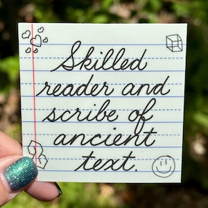 May include: A small, lined paper note with the words "Skilled reader and scribe of ancient text" in elegant cursive script. The note has small doodles of hearts, a cube, a hexagon, and a smiley face.