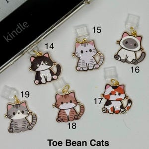May include: A collection of cat-themed charms in various colors and patterns, including black and white, gray, and calico. Each charm has a gold-colored loop and a clear plastic attachment. The text "Toe Bean Cats" is visible at the bottom.