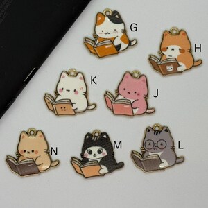 May include: A collection of colorful cat charms, each reading a book. The cats are in various colors, including white, orange, pink, and gray, with gold-colored loops for attaching to jewelry or other crafts. The books are open and have a brown or pink cover.
