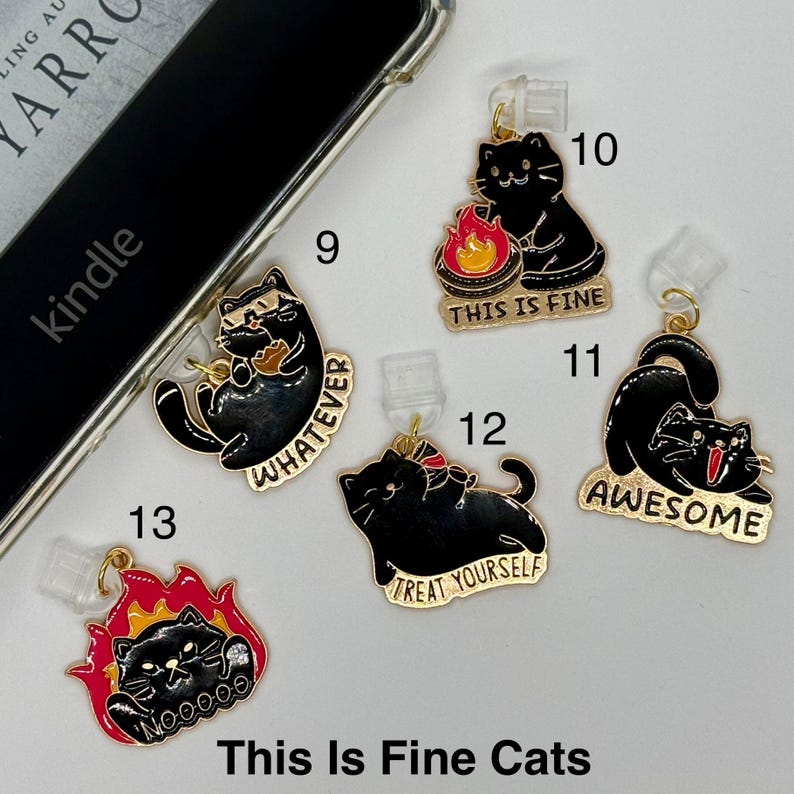 May include: A collection of black cat charms with gold accents, each featuring a different expression and phrase. Phrases include "Whatever," "This is Fine," "Awesome," and "Treat Yourself." The charms are attached to clear phone accessories.