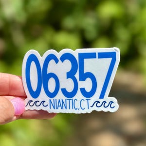 May include: A white sticker with blue text and a wavy border. The sticker displays the numbers "06357" and the text "NIANTIC, CT". The sticker is held up against a blurred green background.