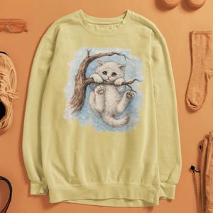 May include: A pale yellow sweatshirt with a graphic of a white kitten clinging to a tree branch. The kitten has blue eyes and is set against a light blue and white backdrop. The sweatshirt is a casual style.