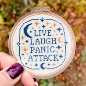 May include: A round sticker with a cross-stitch design. The sticker features the words "LIVE. LAUGH. PANIC. ATTACK." in a blue and orange color scheme, with crescent moons and stars. The sticker is held by a hand.