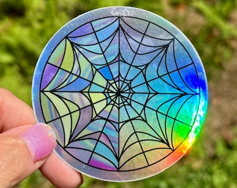 May include: A circular sticker featuring a black spider web design printed on a holographic background that reflects various colors including blue, green, and purple. The web pattern consists of radiating lines and concentric arcs.