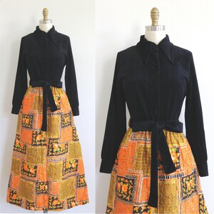 May include: A vintage black velvet dress with long sleeves and a collared neckline. The dress features a patterned skirt with orange, yellow, and black floral squares. A black velvet belt cinches the waist. The dress is displayed on a mannequin.