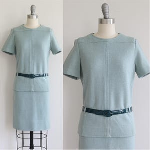May include: A light blue short-sleeved dress featuring a round neckline, vertical seam detailing on the bodice, and a dark teal patent leather belt with an oval buckle at the waist. The dress has a straight skirt silhouette and a smooth texture.