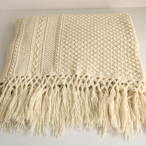 May include: Cream-colored knitted throw blanket with a textured design and fringe. The blanket features a cable knit pattern, a bobble stitch, and a decorative fringe along the bottom edge. The neutral color and intricate details make it a cozy home decor item.