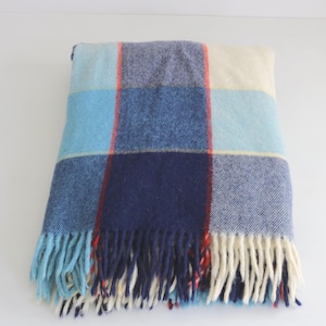 May include: A folded blanket with a checkered pattern in shades of blue, cream, and red. The blanket has a fringed edge with blue and cream tassels. The blanket is made of a soft, woven material.