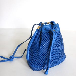 Może przedstawiać: Kobaltowa niebieska torba ze sznurkiem z siatki z pasującym niebieskim skórzanym wykończeniem i sznurkiem. Torba ma złoty metalowy akcent na górze i wiązane zamknięcie na sznurek. Torba jest w stylu vintage.