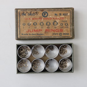May include: A box of jump rings, with a vintage appearance. The box is divided into eight compartments, each holding a quantity of gold-coloured jump rings. The lid features text indicating the product is 14-K white gold filled.