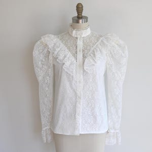 Puede incluir: Blusa de encaje blanco con cuello alto abotonado, detalles de volantes y mangas largas y voluminosas. El patrón floral de encaje es visible en toda la prenda. La blusa tiene una forma entallada y evoca un estilo vintage.