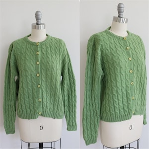May include: A light green cable knit cardigan featuring a crew neckline, long sleeves, and a front button closure with seven matching round buttons. The garment is constructed with a vertical cable pattern throughout the body and sleeves.