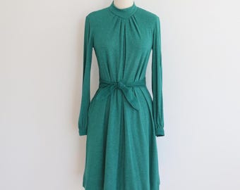 May include: A teal green dress with long sleeves and a high neckline. The dress features a tie-waist detail and a knee-length skirt. The dress is displayed on a mannequin against a white background, showcasing its vintage style.