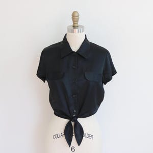 May include: Black short-sleeved button-up shirt with two chest pockets. The shirt is tied at the waist, creating a cropped style. The shirt is displayed on a mannequin against a white background.