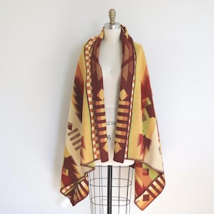 May include: A colorful, patterned blanket draped over a mannequin. The blanket features geometric designs in shades of yellow, brown, orange, and green. The blanket is made of a soft material and has a fringed edge. The blanket is a warm and cozy accessory.