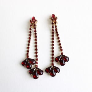 May include: A pair of vintage dangle earrings with a gold-tone setting and deep red rhinestones. The earrings feature a star-shaped top, a chain of small rhinestones, and a three-petal design at the bottom. The earrings are set against a white background.