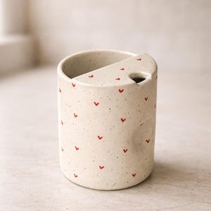Peut inclure: Un r&eacute;cipient en c&eacute;ramique mouchet&eacute;e, de forme cylindrique, avec une section d&eacute;coup&eacute;e sur le dessus. Il est orn&eacute; de petits motifs en forme de c&oelig;ur rouge. Cet article est probablement un porte-brosse &agrave; dents ou un accessoire de salle de bain similaire.