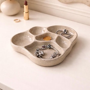 May include: A speckled beige ceramic jewelry tray with multiple compartments. The tray holds a gold coin, a silver bracelet, and a silver chain bracelet. A gold ring and lipstick are visible in the background.