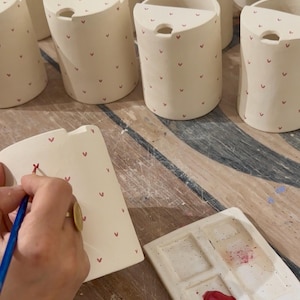 Peut inclure: Plusieurs mugs en c&eacute;ramique blanc cass&eacute; orn&eacute;s de c&oelig;urs rouges. Chaque mug a une forme incurv&eacute;e et une petite ouverture en haut. Une main peint un mug avec un motif de c&oelig;ur rouge &agrave; l'aide d'un pinceau. Une palette de peinture en c&eacute;ramique est &eacute;galement visible.