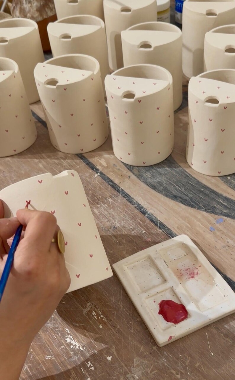 Peut inclure: Plusieurs mugs en c&eacute;ramique blanc cass&eacute; orn&eacute;s de c&oelig;urs rouges. Chaque mug a une forme incurv&eacute;e et une petite ouverture en haut. Une main peint un mug avec un motif de c&oelig;ur rouge &agrave; l'aide d'un pinceau. Une palette de peinture en c&eacute;ramique est &eacute;galement visible.