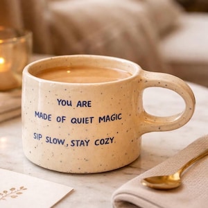 May include: A speckled beige ceramic mug filled with a warm beverage. The mug has a large handle and blue text that reads "YOU ARE MADE OF QUIET MAGIC SIP SLOW, STAY COZY." A gold spoon rests on a folded beige napkin.