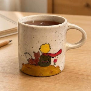 May include: A speckled off-white ceramic mug featuring a hand-painted illustration of a small figure with yellow hair, a red scarf, and a fox sitting on a yellow planet surface next to a small pink rose. The mug has a curved handle and a cylindrical body.