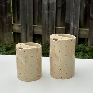 Peut inclure: Deux tasses de voyage en c&eacute;ramique avec couvercles. Les tasses sont beiges avec de petits motifs en forme de c&oelig;ur rouge et un motif mouchet&eacute;. Les tasses sont cylindriques et ont une petite ouverture dans le couvercle pour boire.