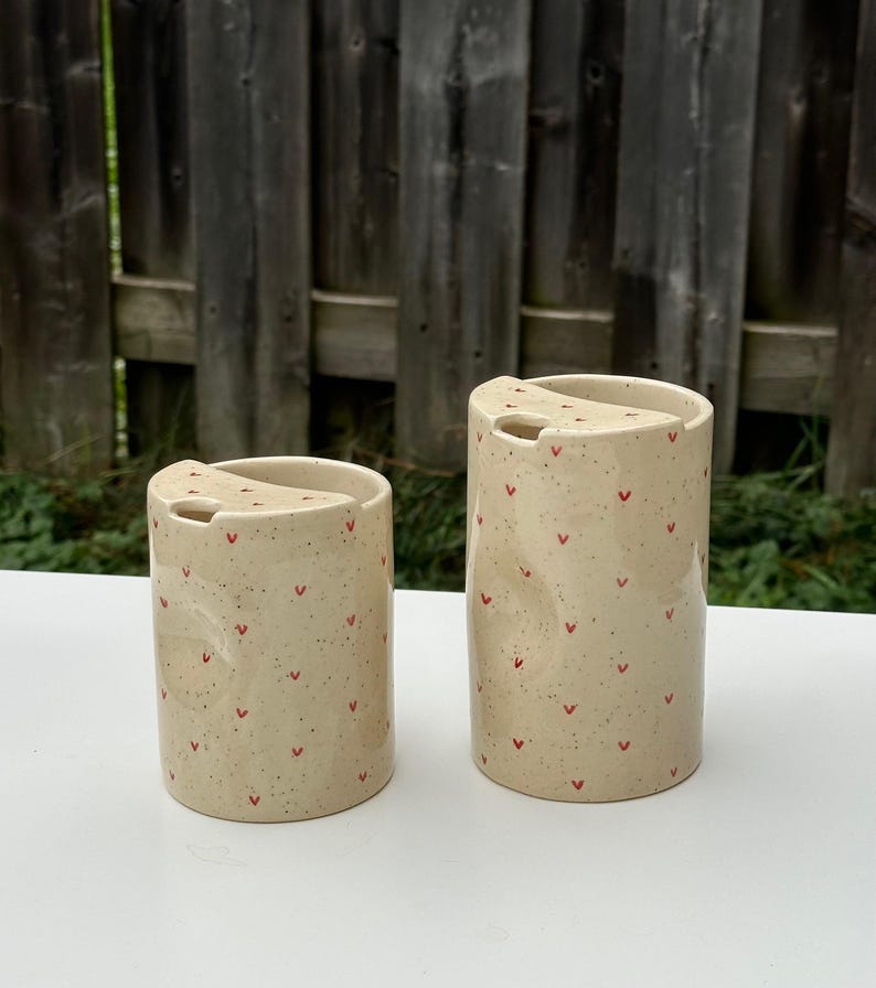 Peut inclure: Deux tasses de voyage en c&eacute;ramique avec couvercles. Les tasses sont beiges avec de petits motifs en forme de c&oelig;ur rouge et un motif mouchet&eacute;. Les tasses sont cylindriques et ont une petite ouverture dans le couvercle pour boire.