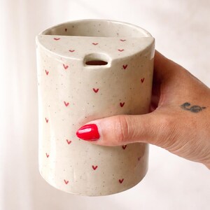 Peut inclure: Un vase cylindrique en c&eacute;ramique avec une finition beige mouchet&eacute;e, orn&eacute; de petits motifs en forme de c&oelig;ur rouge. Le vase a une ouverture rectangulaire en haut et un petit trou. Un objet d&eacute;coratif pour la maison.