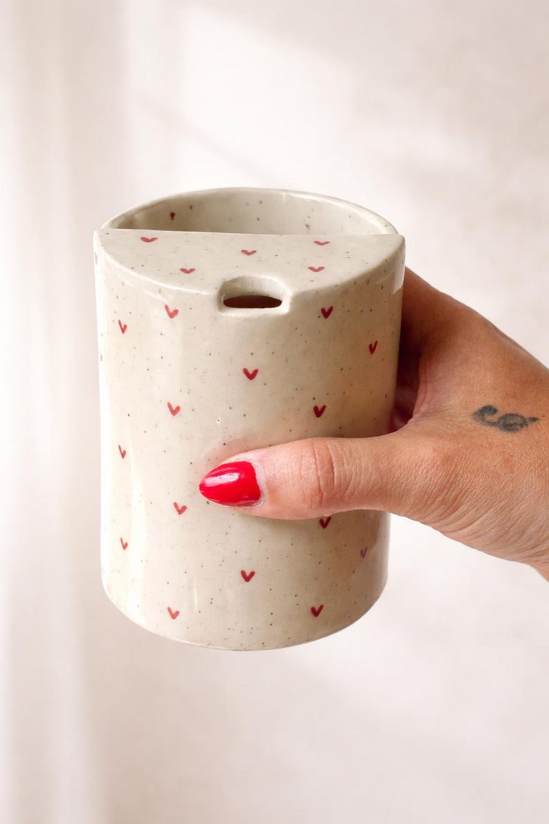 Peut inclure: Un vase cylindrique en c&eacute;ramique avec une finition beige mouchet&eacute;e, orn&eacute; de petits motifs en forme de c&oelig;ur rouge. Le vase a une ouverture rectangulaire en haut et un petit trou. Un objet d&eacute;coratif pour la maison.