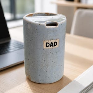 May include: A pale blue ceramic tumbler with the word "DAD" on a small label. The tumbler is cylindrical with a small opening at the top. It sits on a wooden desk next to a notebook and pen.