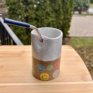 May include: A white ceramic mug with a black handle and the text "But First, Coffee" in a bold, black font. The mug is set against a blurred background, suggesting a cosy setting.