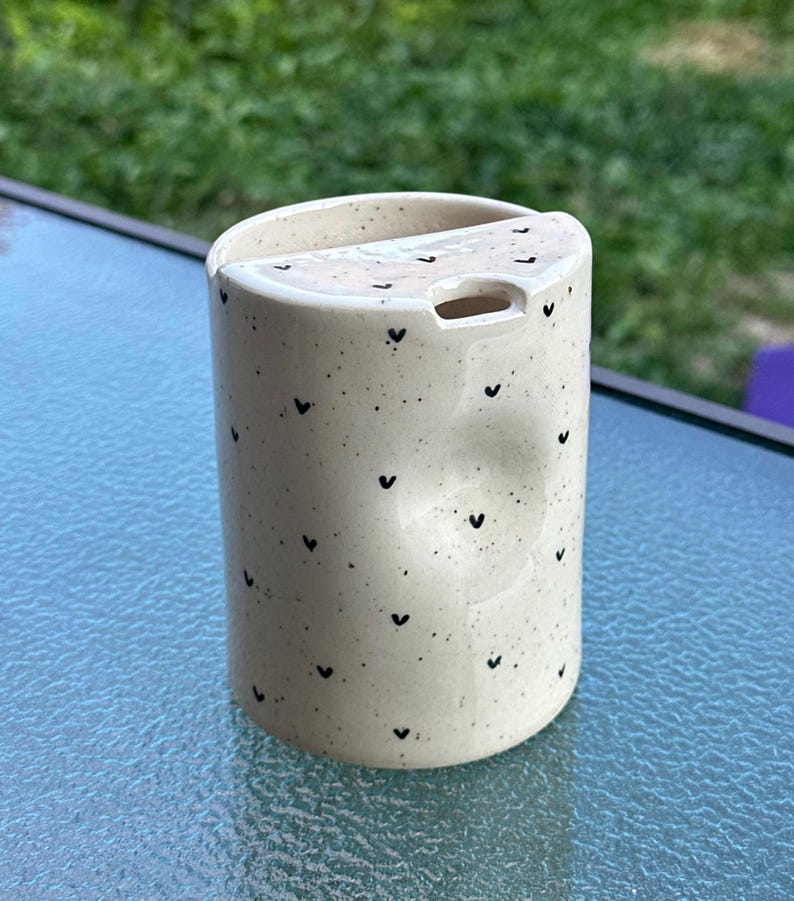 Peut inclure: Une tasse en c&eacute;ramique blanche avec une anse noire et le texte "But First, Coffee" en noir, dans une police manuscrite. La tasse est pos&eacute;e sur une surface blanche.