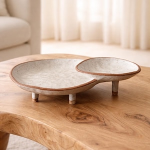 May include: A speckled white ceramic serving dish with a connected smaller dish. The rim is a warm brown colour. The larger dish has three short legs, and the smaller dish has two. The dish is on a wooden table.
