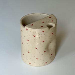Peut inclure: Une tasse en c&eacute;ramique tachet&eacute;e avec un repose-cuill&egrave;re int&eacute;gr&eacute;. La tasse blanc cass&eacute; est orn&eacute;e de petits c&oelig;urs rouges et de mouchetures noires. Le repose-cuill&egrave;re est de couleur et de forme assorties, avec un trou pour le manche de la cuill&egrave;re.