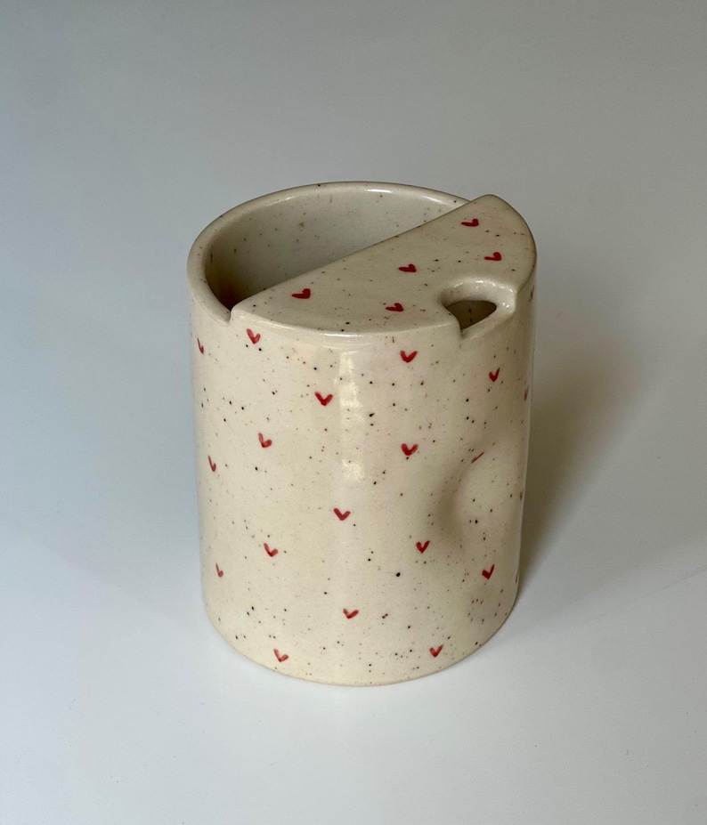 Peut inclure: Une tasse en c&eacute;ramique tachet&eacute;e avec un repose-cuill&egrave;re int&eacute;gr&eacute;. La tasse blanc cass&eacute; est orn&eacute;e de petits c&oelig;urs rouges et de mouchetures noires. Le repose-cuill&egrave;re est de couleur et de forme assorties, avec un trou pour le manche de la cuill&egrave;re.