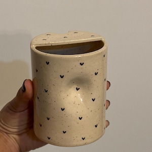Peut inclure: Un porte-brosse &agrave; dents cylindrique en c&eacute;ramique avec une finition beige mouchet&eacute;e et de petits motifs en forme de c&oelig;ur noirs. Le support a une ouverture incurv&eacute;e en haut et une petite indentation sur le c&ocirc;t&eacute;.
