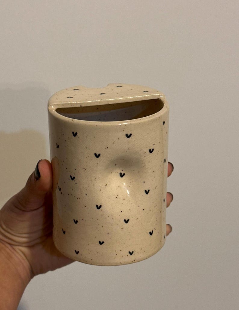 Peut inclure: Un porte-brosse &agrave; dents cylindrique en c&eacute;ramique avec une finition beige mouchet&eacute;e et de petits motifs en forme de c&oelig;ur noirs. Le support a une ouverture incurv&eacute;e en haut et une petite indentation sur le c&ocirc;t&eacute;.