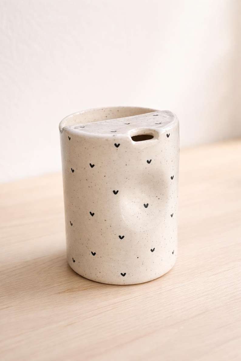 Peut inclure: Une tasse en c&eacute;ramique blanche avec une anse noire et l'inscription "But First, Coffee" en gras noir. La tasse est pos&eacute;e sur un fond flou, sugg&eacute;rant un cadre chaleureux.