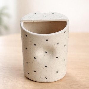 Peut inclure: Un porte-objet en c&eacute;ramique mouchet&eacute;e avec un dessus arrondi et une ouverture incurv&eacute;e. La surface blanc cass&eacute; est orn&eacute;e de petits motifs noirs en forme de c&oelig;ur. Le support est cylindrique et repose sur une surface en bois clair.