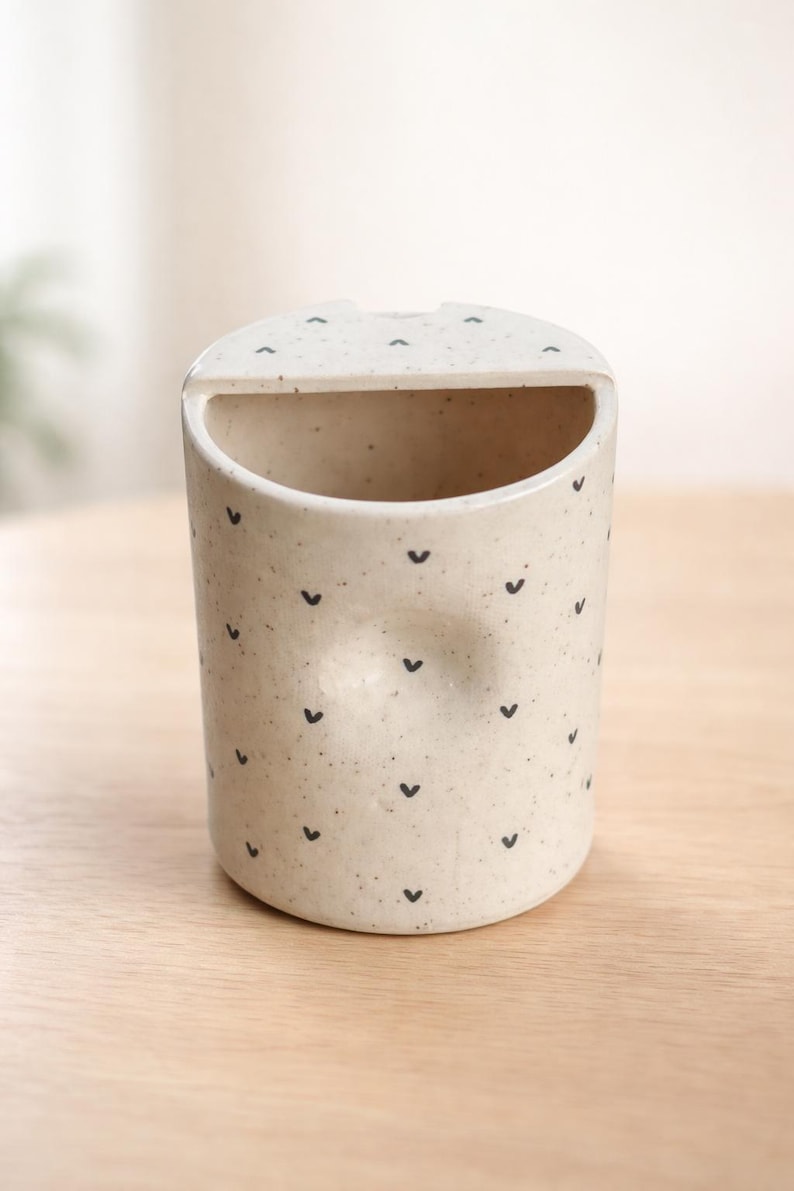 Peut inclure: Un porte-objet en c&eacute;ramique mouchet&eacute;e avec un dessus arrondi et une ouverture incurv&eacute;e. La surface blanc cass&eacute; est orn&eacute;e de petits motifs noirs en forme de c&oelig;ur. Le support est cylindrique et repose sur une surface en bois clair.