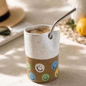 Peut inclure: Un gobelet cylindrique en céramique avec une moitié supérieure blanche tachetée et une moitié inférieure brune ornée de visages souriants peints. Le gobelet contient une boisson froide avec des glaçons et une paille en métal argenté.