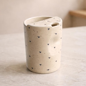 Peut inclure: Un gobelet en c&eacute;ramique cr&egrave;me avec un dessus asym&eacute;trique unique et une petite ouverture. Le gobelet est parsem&eacute; de minuscules motifs en forme de c&oelig;ur bleu fonc&eacute;. Le design est minimaliste et moderne, adapt&eacute; aux boissons.