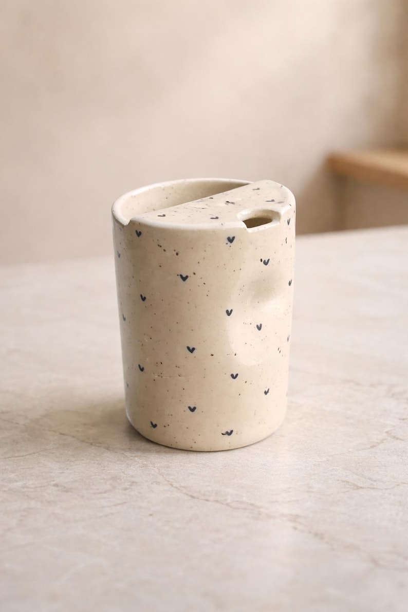 Peut inclure: Un gobelet en c&eacute;ramique cr&egrave;me avec un dessus asym&eacute;trique unique et une petite ouverture. Le gobelet est parsem&eacute; de minuscules motifs en forme de c&oelig;ur bleu fonc&eacute;. Le design est minimaliste et moderne, adapt&eacute; aux boissons.