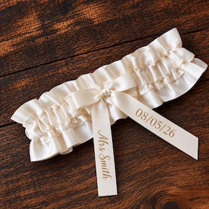 May include: An ivory satin garter featuring a ruffled edge and a central bow. Two satin ribbons hang from the bow, with the text 'Mrs Smith' printed in gold on one ribbon and the date '08/05/26' printed in gold on the other ribbon.