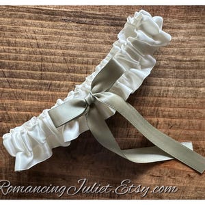 May include: A white garter featuring a gathered, ruffled fabric band and a centered sage green satin ribbon tied into a bow with two long, trailing ends. The accessory has a smooth, lustrous finish and is displayed against a textured wood surface.